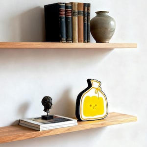 May include: Two wooden shelves against a white wall. The top shelf holds a collection of books and a gray vase. The bottom shelf displays a small bust, a stack of books, and a yellow and black decorative item with a smiling face.