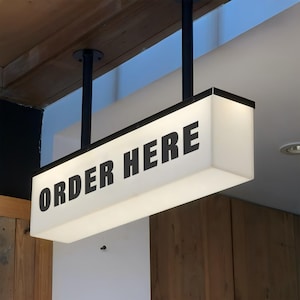以下が含まれることがあります： 太字の黒文字で「ORDER HERE」と書かれた長方形の照明付き看板。看板は2本の黒いポールで天井から吊り下げられています。白い光る表面と黒い縁があります。