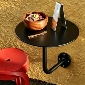 Könnte beinhalten: Ein schwarzer, runder, an der Wand befestigter Tisch mit einer Holzschüssel mit Essen, einem Metallschild mit der Nummer 1 und einer goldenen Gabel. Ein roter Hocker befindet sich im Vordergrund. Der Tisch wird von einem schwarzen, gebogenen Metallarm getragen.