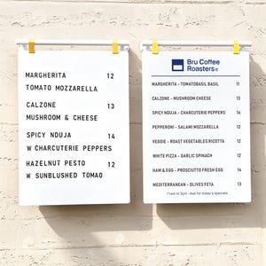 May include: Two white rectangular menu boards with black lettering suspended by white bars and yellow clips. The left board lists pizza options like Margherita and Calzone. The right board features the Bru Coffee Roasters logo and menu items.