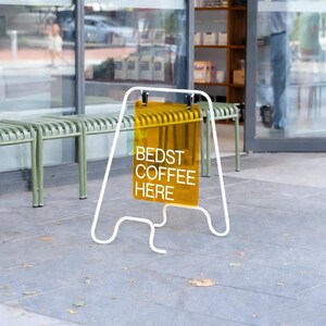 May include: A-frame sign with the text "BEDST COFFEE HERE" in white on a yellow translucent panel. The sign is supported by a white metal frame and sits on a gray concrete surface. The background includes a storefront with benches.