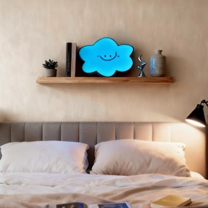 May include: A blue cloud-shaped night light with a smiling face sits on a wooden shelf. The shelf also holds a small potted plant, a book, a decorative sculpture, and a vase. The light is above a bed with white pillows and a grey headboard.