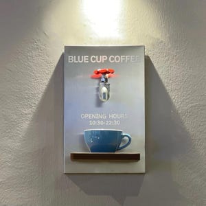 Puede incluir: Un letrero mural para "BLUE CUP COFFEE" con una taza de café azul sobre una repisa de madera. Encima de la taza hay un grifo plateado con un mango rojo. El letrero también muestra el horario de apertura: 10:30-22:30.