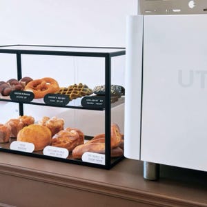 May include: A black metal two-tier display case filled with various golden-brown baked goods including pretzels, waffles, and pastries. Each item is labeled with a small black sign. Next to the case stands a large, plain white appliance.
