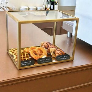 Könnte beinhalten: Eine Glasvitrine mit goldenem Rahmen enthält eine Waffel, eine Brezel und mehrere Croissants. Vor dem Gebäck befinden sich drei schwarze Schilder mit der Aufschrift "CROISSANT $2 FRESH TODAY", "COFFEE & WALNUT CAKE $7.00" und "COFFEE & WALNUT CAKE $7.00".