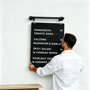 Puede incluir: Una pizarra negra con texto blanco, colgada en una pared blanca. El menú enumera opciones de pizza como Margherita, Calzone y Spicy Salami. Una persona con una camisa blanca sostiene el menú.