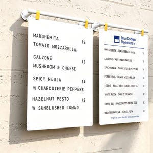 May include: Two white rectangular menu boards hanging from a white horizontal bar with yellow clips. The left board lists Margherita, Calzone, Spicy Nduja, and Hazelnut Pesto. The right board features the Bru Coffee Roasters logo and pizza options.
