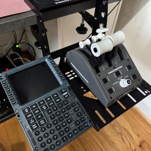 May include: A flight simulator setup featuring a gray control panel with numerous buttons and a display screen, alongside a throttle quadrant and a black mounting bracket. The setup is designed for flight simulation enthusiasts.