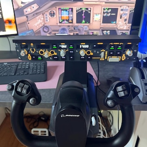 May include: Black flight simulator yoke with the Boeing logo, and a control panel with illuminated gauges and dials. The simulator setup includes a keyboard and a monitor displaying a cockpit view.