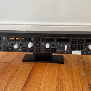 May include: A black flight simulator panel with various buttons, knobs, and switches. The panel is mounted on a black stand and sits on a wooden surface. The panel has white and red accents.