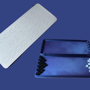 Könnte beinhalten: Ein weißes rechteckiges Spielbrett mit dem Wort "HAPPY" und fischförmigen Designs. Das Brett befindet sich neben einem offenen, rechteckigen Spielkoffer mit schwarzen und weißen Spielsteinen auf einer blauen Oberfläche.