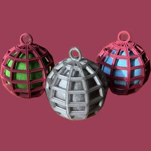 Könnte beinhalten: Drei dekorative Kugelornamente. Eines ist silberfarben, die anderen beiden sind rot mit grünen und blauen Innenräumen. Jedes hat ein geometrisches Käfigdesign und eine kleine Schlaufe zum Aufhängen. Der Hintergrund ist weinrot.