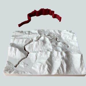 May include: A white 3D topographical model with a winding river carved through the landscape. A separate, red, ribbon-like object hovers above the model, possibly representing a road or path. The model is set against a light gray background.