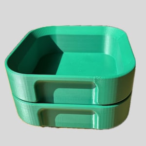 Può includere: Due vassoi quadrati impilati, color teal. Ogni vassoio ha angoli arrotondati e una maniglia incassata sul davanti. I vassoi sono realizzati in un materiale solido e sembrano stampati in 3D.