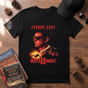 May include: Black t-shirt featuring a portrait of Johnny Cage from Mortal Kombat II. The design includes the text "JOHNNY CAGE" and "MORTAL KOMBAT II" in red. The shirt is paired with a black cap and sunglasses.