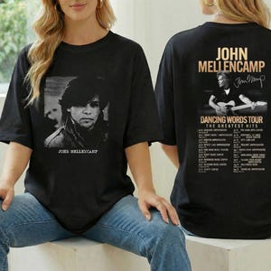 May include: Black t-shirt featuring a black and white image of John Mellencamp on the front. The back of the shirt displays a photo of Mellencamp playing guitar, the text "John Mellencamp" and "Dancing Words Tour The Greatest Hits".