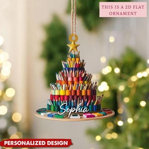 May include: A 2D flat hanging ornament shaped like a Christmas tree made of stacked colorful pencils and paint pots. The name Sophia is written in white script across the base. A gold star sits at the top, suspended by a twine string.