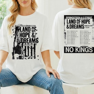 Puede incluir: Camiseta blanca con un gráfico en blanco y negro de un músico y el texto "LAND OF HOPE & DREAMS AMERICAN TOUR" en la parte delantera. La parte trasera de la camiseta tiene un calendario de gira y las palabras "NO KINGS".