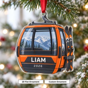 Könnte beinhalten: Ein flacher, transparenter Acryl-Weihnachtsbaumschmuck in Form einer orangefarbenen Skigondel mit rotem Band. Die Gondel zeigt eine Berglandschaft, den Namen LIAM und das Jahr 2026 in Weiß sowie zwei seitlich befestigte Skier.