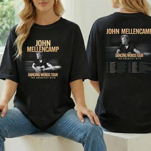 May include: Black t-shirt featuring John Mellencamp's name and image, with "Dancing Words Tour The Greatest Hits" text. The back of the shirt also displays the tour name, image, and tour dates. The shirt is worn by a person.