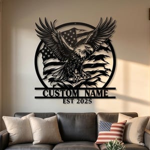 May include: A black metal wall art piece featuring a circular frame with a detailed eagle in flight over a stylized American flag. Below the circle, a horizontal bar displays the text CUSTOM NAME and the year EST 2025 in bold lettering.