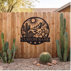May include: A circular black metal wall sign featuring a desert landscape with a sun, mountains, and various cacti. Below the circular design is a banner with the text CUSTOM NAME and three star cutouts centered at the bottom of the metal piece.