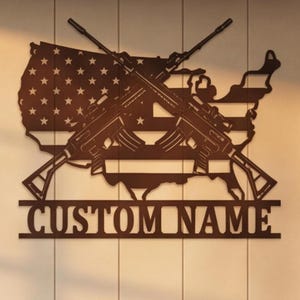 Puede incluir: Un letrero de pared de metal marrón oscuro que presenta una silueta del mapa de los Estados Unidos integrado con el patrón de la bandera estadounidense, dos rifles cruzados sobre el mapa y una barra horizontal con el texto CUSTOM NAME.
