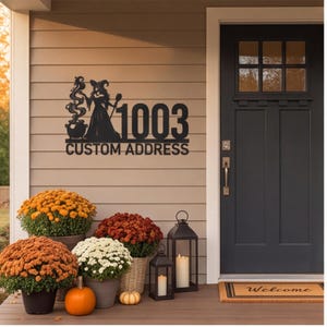 May include: A black metal wall sign featuring a silhouette of a witch standing next to a bubbling cauldron, the number 1003, and the text CUSTOM ADDRESS. The sign is mounted on a beige horizontal siding wall next to a dark gray front door.