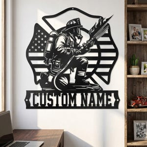 Puede incluir: Arte de pared de metal negro que representa a un bombero arrodillado sosteniendo una manguera con llamas. El diseño incluye una bandera estadounidense y una pancarta que dice "CUSTOM NAME". La obra de arte está montada en una pared blanca.