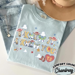 May include: A light blue t-shirt featuring the text I KNEW IT, I KNEW YOU in a multi-colored font with patterns inspired by cowboy and space characters. The design includes small stars, clouds, a rocket, and a red heart with the letters TS inside.