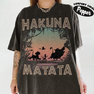 May include: Dark grey t-shirt with the words "HAKUNA MATATA" in a distressed font. The shirt features a colourful graphic of silhouettes of Simba, Timon, Pumbaa, and a bird. The brand name "Comfort Colors Pepper" is printed on the upper right.