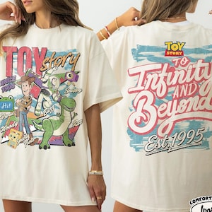 Pode incluir: Uma t-shirt cor de marfim com gr&aacute;ficos de Toy Story. A frente mostra Woody, Buzz Lightyear, Rex e texto incluindo "Toy Story", "Since 1995" e "Smarty Pants". As costas apresentam o log&oacute;tipo de Toy Story e o texto "To Infinity and Beyond Est. 1995".