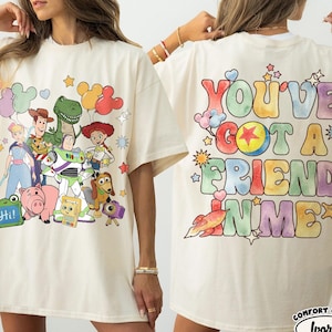 May include: An ivory t-shirt featuring a graphic print of Toy Story characters on the front and the text "YOU'VE GOT A FRIEND IN ME" in colourful, bubble-style lettering on the back, surrounded by small star, heart, and rocket illustrations.