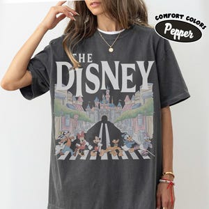 Może przedstawiać: Ciemnoszara koszulka z napisem "THE DISNEY" dużymi białymi literami. Grafika przedstawia postacie Disneya przechodzące przez ulicę, z zamkiem w tle. Koszulka ma wygląd vintage, a słowa "Comfort Colors Pepper" są w czarnym owalu.