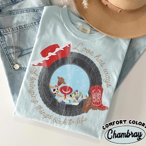 May include: A light blue t-shirt featuring a graphic of a black tire framing cartoon characters, a red cowboy hat, and a red cowboy boot. The text reads Love has ways of bringing things back to life. The shirt is placed near blue jeans and a tan hat.