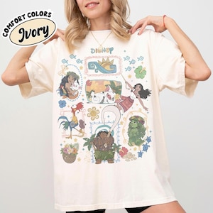 May include: Ivory-coloured t-shirt featuring Disney characters from the movie Moana. The shirt has a crew neck and short sleeves. The graphic includes illustrations of Moana, Maui, Hei Hei, Pua, and other characters. The text "Disney" is printed at the top.
