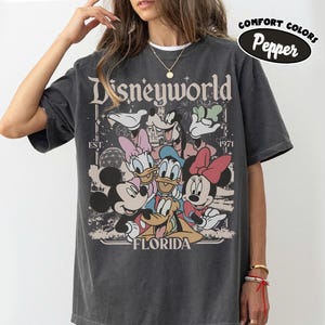 May include: Dark gray t-shirt featuring a graphic of Disney characters including Mickey, Minnie, Donald, and Daisy Duck. The shirt has the text "Disneyworld Florida" and "EST. 1971". The shirt is labeled "Comfort Colors Pepper".