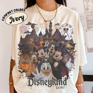 May include: An ivory t-shirt featuring a graphic of Disney characters in Halloween costumes, including Mickey Mouse as a vampire, Minnie Mouse as a witch, and Donald Duck as a ghost, with a castle, ferris wheel, and the text Disneyland Est. 1955.