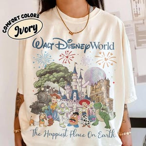 May include: Ivory-colored t-shirt featuring a colorful graphic of Walt Disney World with fireworks and cartoon characters. The text "Walt Disney World" and "The Happiest Place On Earth" are printed on the shirt.