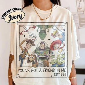 May include: Ivory-colored t-shirt featuring a graphic of Toy Story characters. The graphic includes Woody, Buzz Lightyear, and other characters from the movie. Text on the shirt reads "YOU'VE GOT A FRIEND IN ME" and "EST.1995".