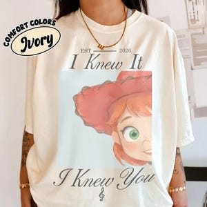 Op de afbeelding: Een ivoorkleurig t-shirt met een afbeelding van een stripfiguur met rood haar en een rode cowboyhoed. De tekst "EST 2026 I Knew It" staat boven de afbeelding en "I Knew You" met een muzieknoot eronder. Een logo toont "COMFORT COLORS IVORY".
