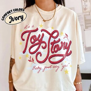 May include: An ivory short-sleeve t-shirt featuring a red rope-textured graphic that reads "it's a Toy Story" with the text "Baby, just say 'yes'" below. The design includes yellow stars, light blue clouds, and small red floral accents.