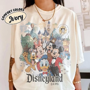 May include: Ivory-colored t-shirt featuring a colorful Disneyland graphic with Mickey Mouse, Minnie Mouse, Donald Duck, and other characters. The shirt has the words "Comfort Colors" and "Ivory" printed on it, and the word "Disneyland" is printed at the bottom.