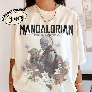 May include: Ivory-colored t-shirt with a graphic featuring The Mandalorian and Grogu surrounded by flowers and butterflies. The text "MANDALORIAN - THIS IS THE WAY" is printed above the image. The shirt has a Comfort Colors label.