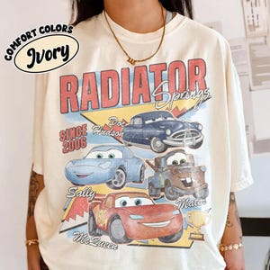 May include: Ivory-colored t-shirt featuring a graphic of characters from the movie Cars, including Doc Hudson, Sally, and Lightning McQueen. The design includes the text "Radiator Springs" and "Since 2006."