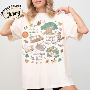 Puede incluir: Camiseta de color marfil con un dise&ntilde;o gr&aacute;fico colorido que presenta personajes de dibujos animados, un &aacute;rbol y las frases "hakuna matata" y "adventure is out there". La camiseta tambi&eacute;n tiene el texto "anyone can be anything".