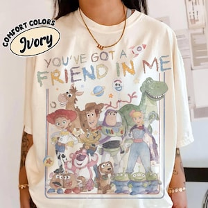 May include: An ivory t-shirt featuring a graphic print of various animated characters from the Toy Story franchise. The text "YOU'VE GOT A FRIEND IN ME" is printed above the characters in a multi-colored, distressed font style.