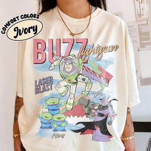 May include: Ivory t-shirt with a graphic of Buzz Lightyear, aliens, and Zurg from Toy Story. The design includes the text "BUZZ lightyear" and "LASER BLAST". The shirt is labelled "COMFORT COLORS" and "Ivory".