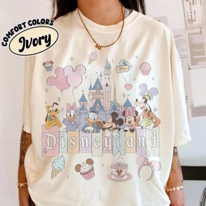 May include: An ivory t-shirt featuring a graphic print of a castle, Mickey Mouse, Minnie Mouse, Donald Duck, Daisy Duck, Goofy, and Pluto. The design includes pastel-colored balloons, cupcakes, a coffee cup, and the text Disneyland in a serif font.