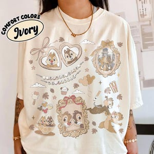 May include: Ivory-colored t-shirt featuring cartoon characters and the text "Disney with my bestie." The shirt has various framed images of cartoon characters, popcorn, and other graphics. The shirt is a Comfort Colors brand.